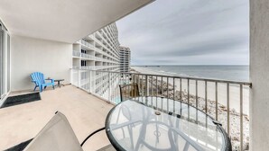 Un balcón con una mesa y sillas de vidrio, una silla azul, una mesa pequeña y una barandilla blanca con vistas a la playa.