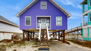 A purple house with a wooden staircase leading to the entrance, situated on stilts over a sandy area. - Centrally-located Gulf front paradise wfurnished sun deck & gorgeous water view (Gulf Shores)