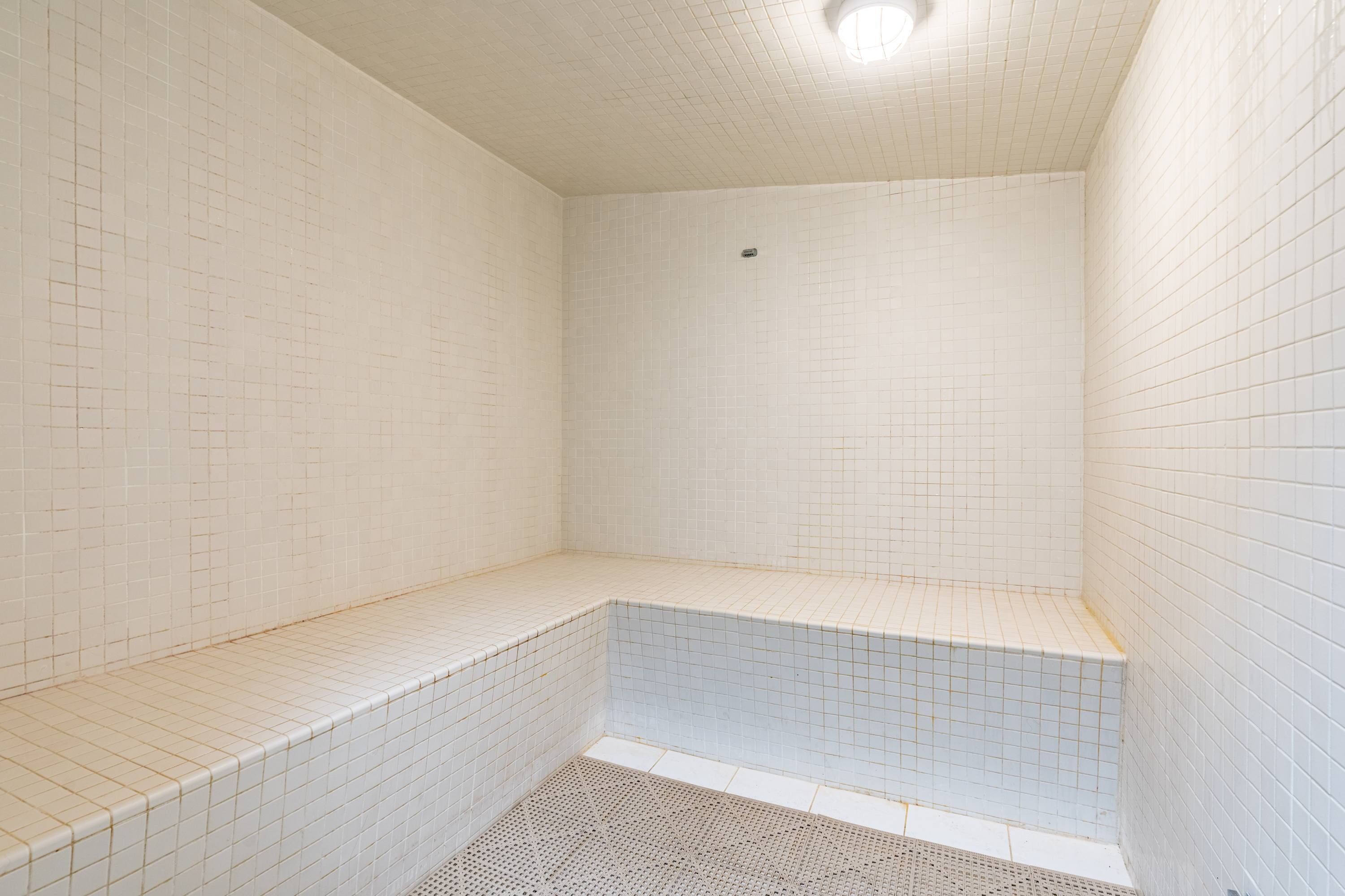 A bathroom with white tiled walls and floor, a bench, and a shower area.