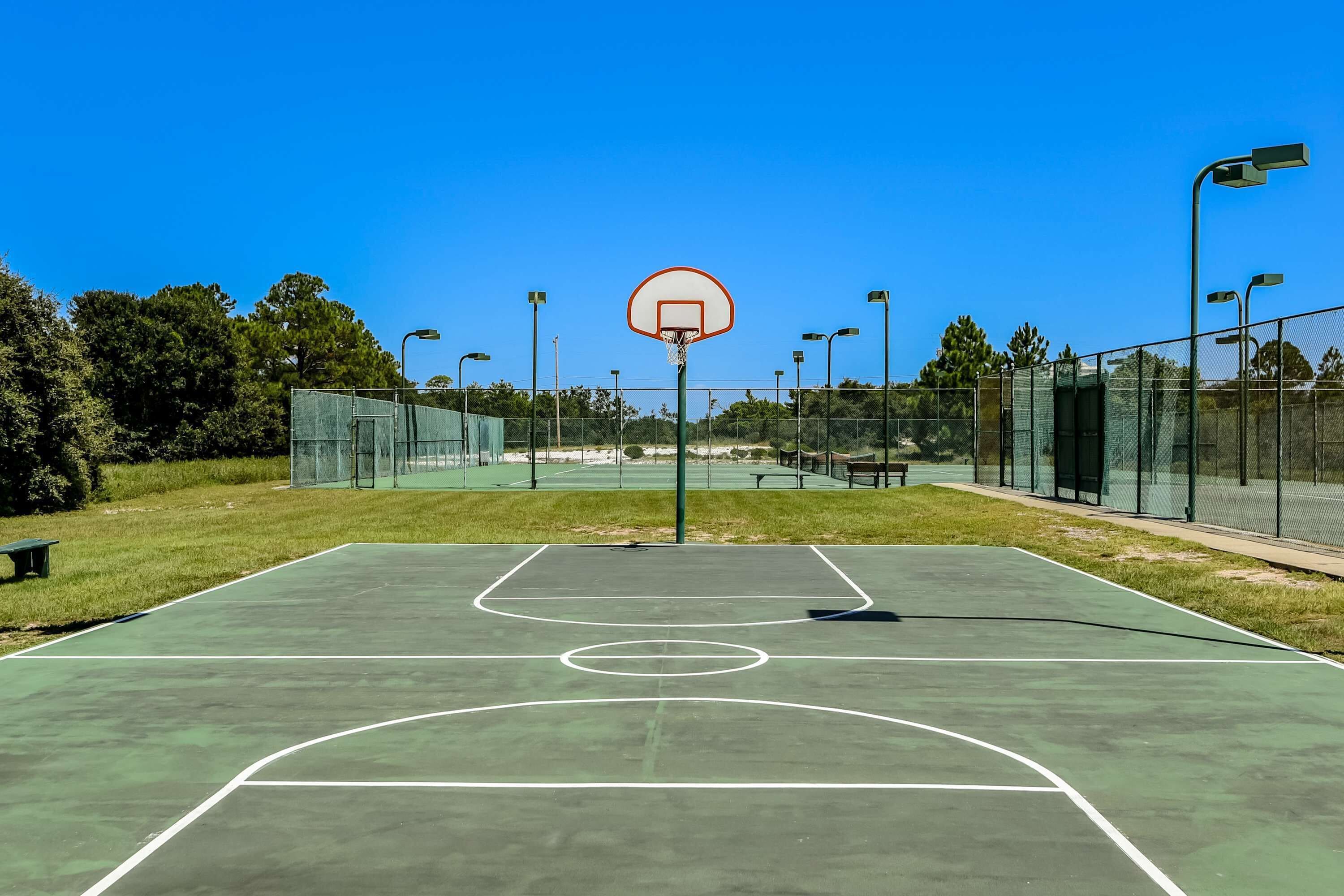 Un terrain de basket en plein air, équipé d’un panier, entouré d’une clôture, de bancs et de lampadaires.