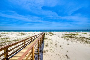 Ein Holzsteg führt zu einem Strand mit Dünen und einem strahlend blauen Himmel.