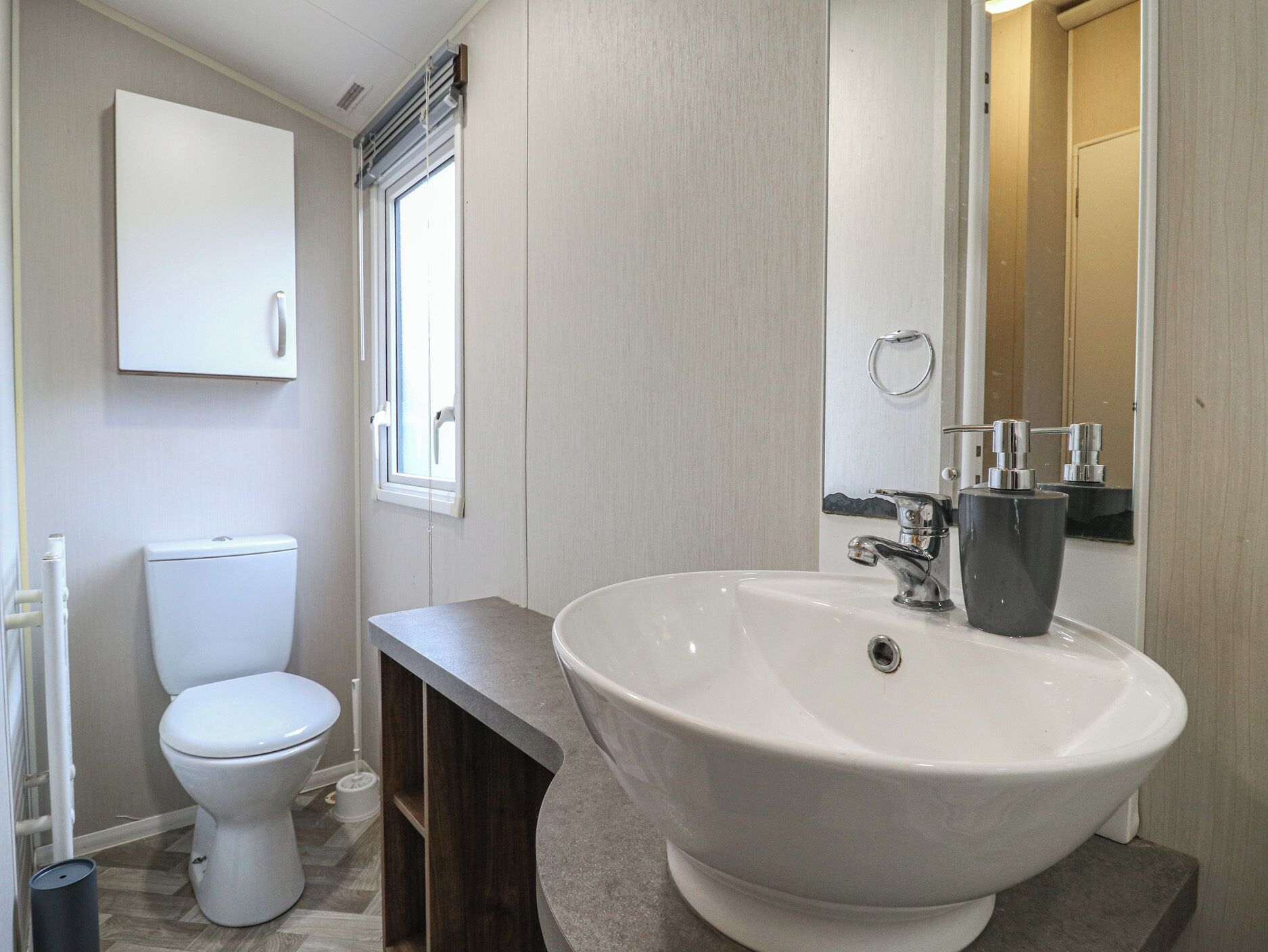 Une salle de bain avec des toilettes blanches, un grand lavabo blanc doté d’un robinet noir, un miroir et une fenêtre avec des stores.
