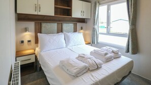 Ein ordentlich bezogenes Bett mit weißen Leinen und Handtüchern, ein Holz-Kopfteil, ein kleiner Nachttisch mit Lampe und ein Fenster mit Vorhängen.