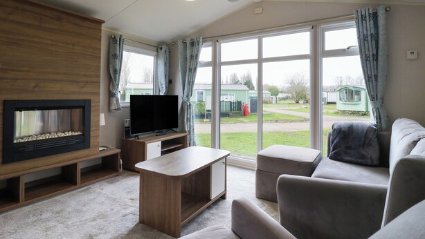 Ein Wohnzimmer mit Flachbildfernseher, einer Holz-Unterhaltungsecke und einem großen Fenster mit Blick auf ein grünes Gebäude und einen Weg.