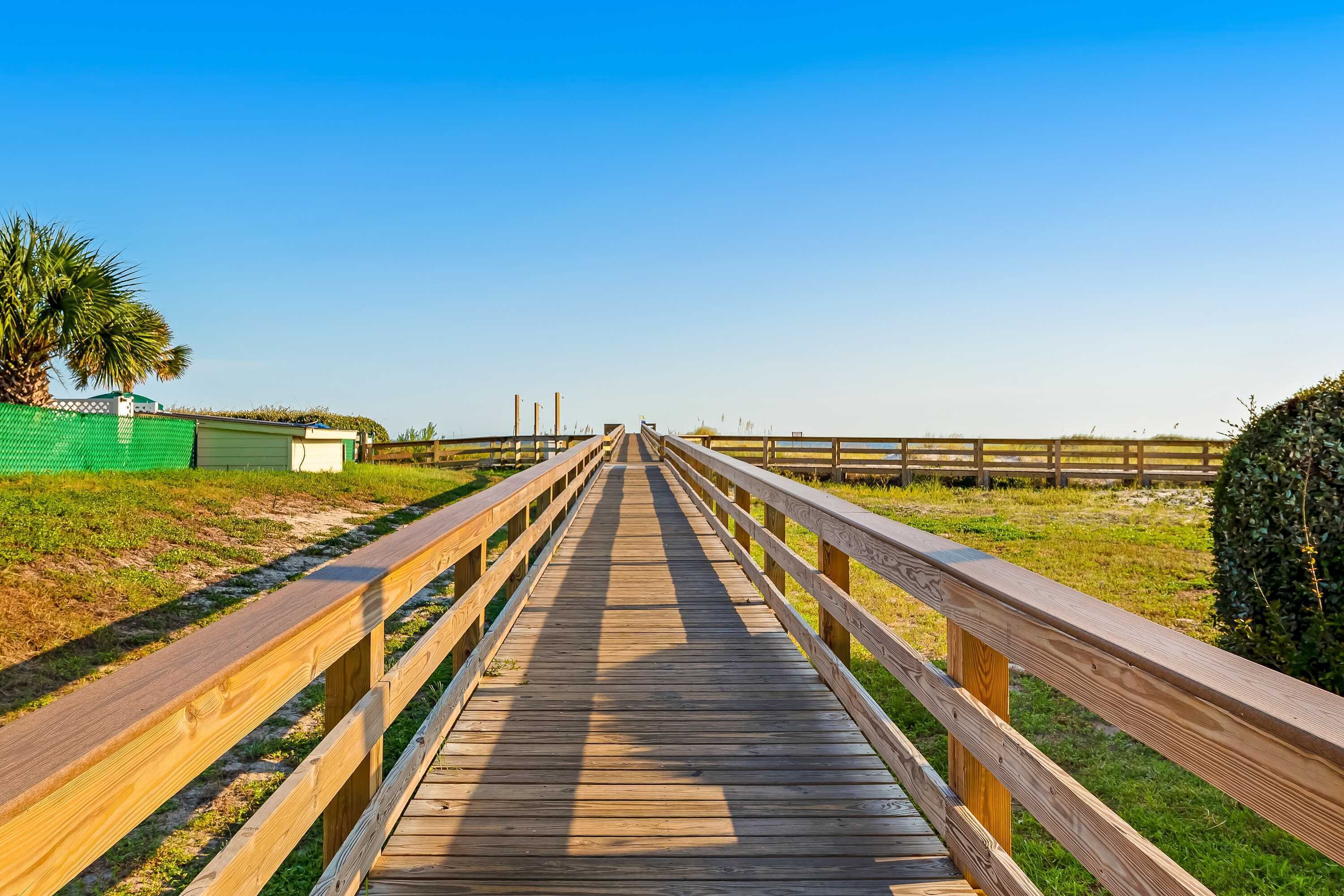 A wooden boardwalk leading to a building with a green roof, surrounded by grass and a fence.