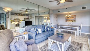 A living room with a sofa, coffee table, and dining area with benches. - Spacious and inviting seaside condo with shared pools, sauna (Fort Morgan)