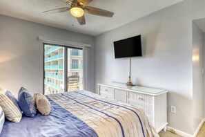 Een slaapkamer met een bed, een plafondventilator, een aan de muur bevestigde televisie, een witte dressoir en een glazen schuifdeur die uitkomt op een balkon.