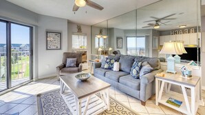 A living room with a sofa, armchair, coffee table, and a side table with a lamp. There is a ceiling fan, a large mirror, and a window with blinds. - Spacious and inviting seaside condo with shared pools, sauna (Fort Morgan)