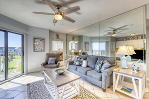 A living room with a sofa, armchair, coffee table, and a side table with a lamp. There is a ceiling fan, a large mirror, and a window with blinds. - Spacious and inviting seaside condo with shared pools, sauna (Fort Morgan)