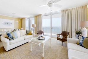 Ein Wohnzimmer mit einem weißen Sofa, einem Glastisch, Korbstühlen, einem Deckenventilator und Meerblick durch große Fenster.