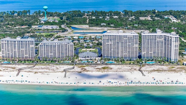 A coastal resort with multiple high-rise buildings, a sandy beach, and a water tower. - Beautiful Home Steps From the Bay with Shared Pool & Hot Tub & Free WiFi, Balcony (Fort Morgan)