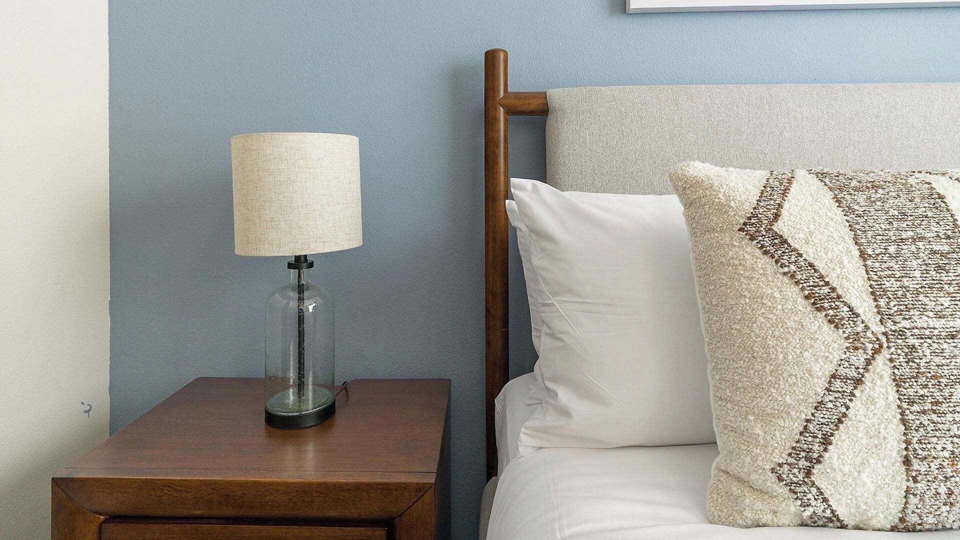 A bed with white bedding and a decorative pillow, a bedside lamp on a wooden nightstand, and a blue wall.