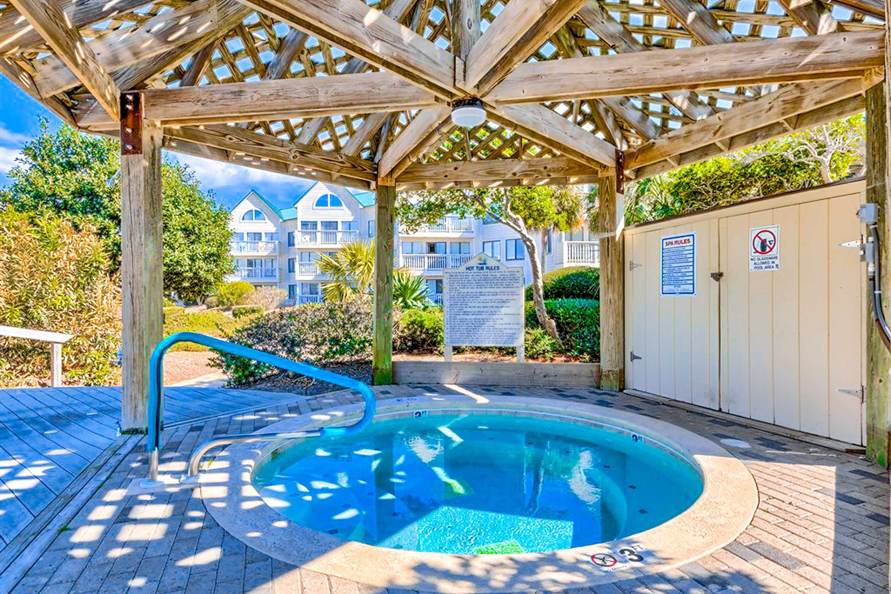 A covered hot tub area with a wooden pergola, a pool, and a building in the background.