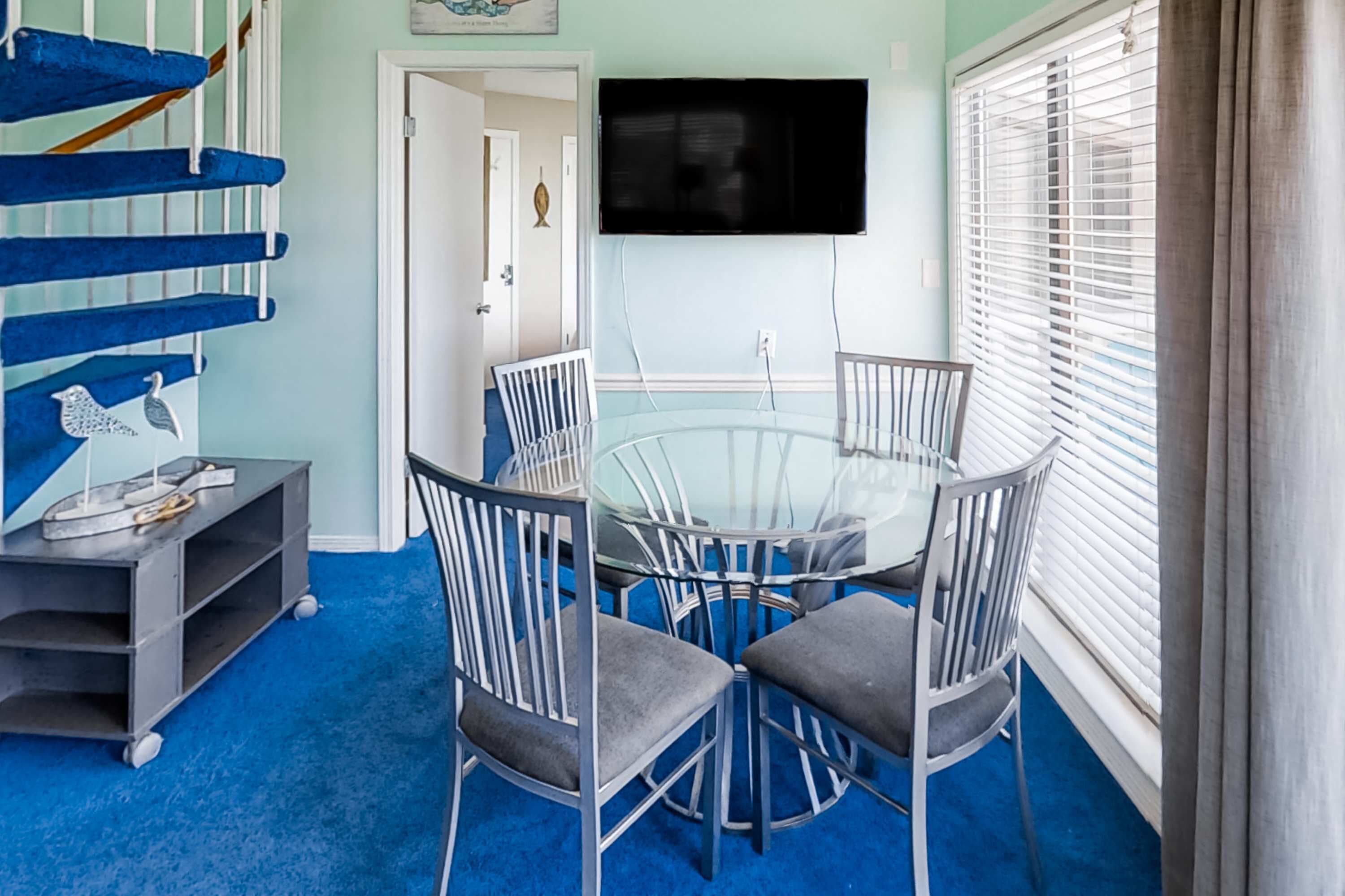 A dining area with a glass table and metal chairs, a blue carpet, a TV mounted on the wall, and a staircase with blue steps.
