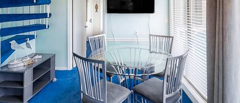 A dining area with a glass table and metal chairs, a blue carpet, a TV mounted on the wall, and a staircase with blue steps.