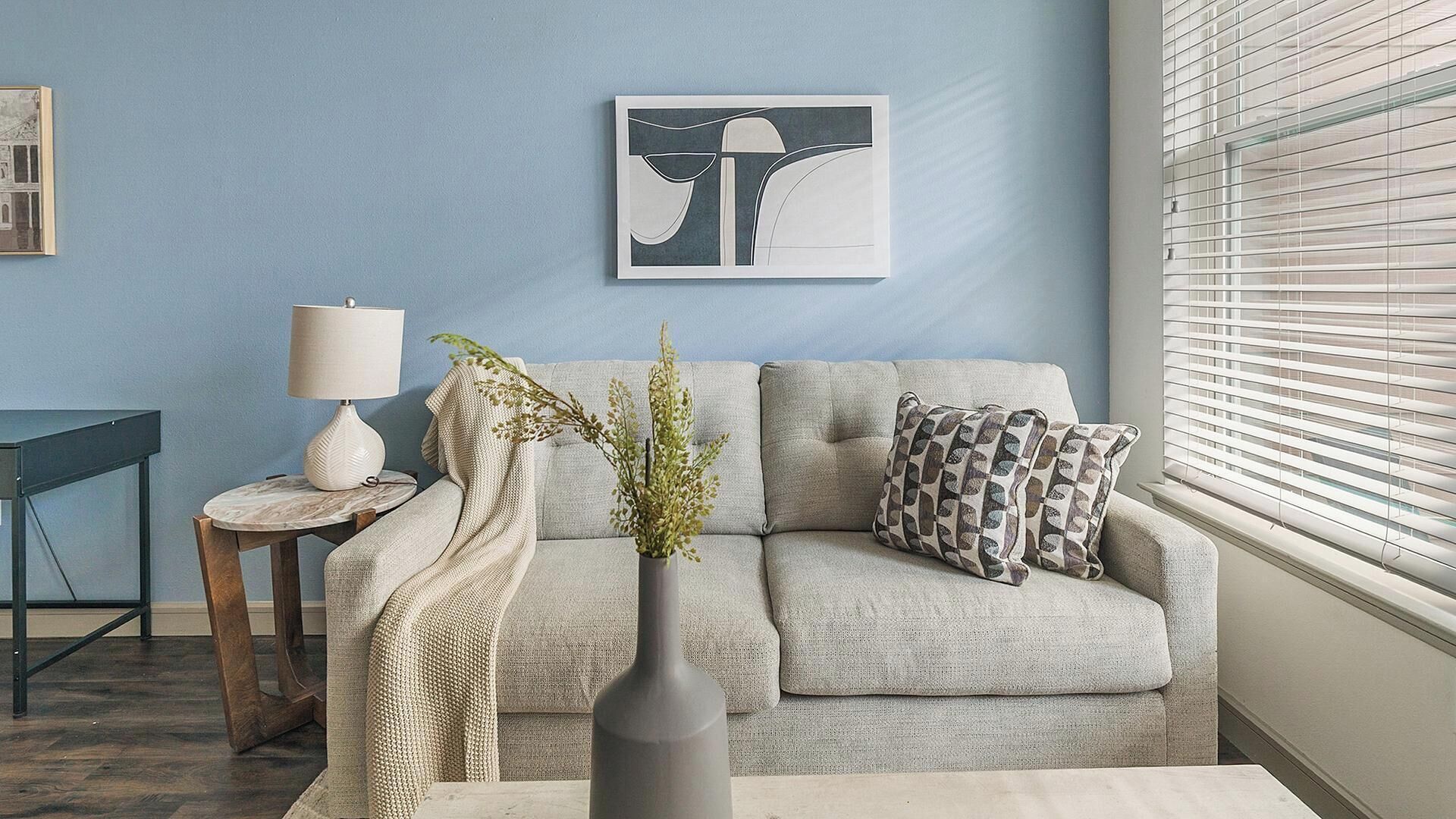 Ein Wohnzimmer mit einer Couch, einem Beistelltisch mit Lampe, einer Vase mit Pflanzen und einem gerahmten Kunstwerk an der Wand.