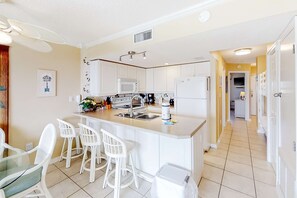 A modern kitchen with white cabinetry, a central island with bar stools, a microwave, and a refrigerator. - Gulf View Condo w Balcony, WiFi, AC & Shared Pools, Hot Tubs, Tennis, Gym (Fort Morgan)