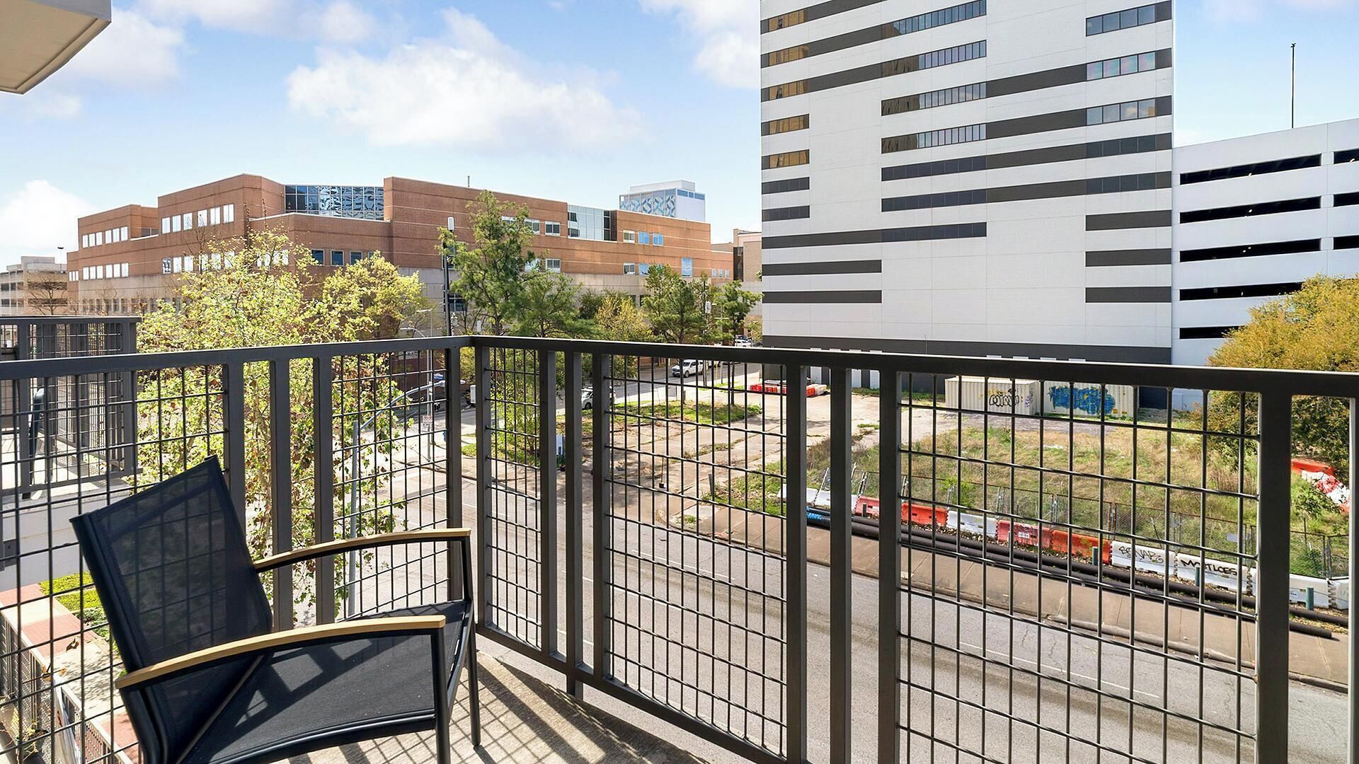 A balcony with a chair, a view of a cityscape with modern buildings, and a street with parked cars.