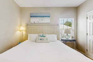 A bedroom with a bed, a nightstand, a lamp, a window with blinds, and a beach-themed pillow. - Gorgeous cottage with great amenties (Fort Morgan)