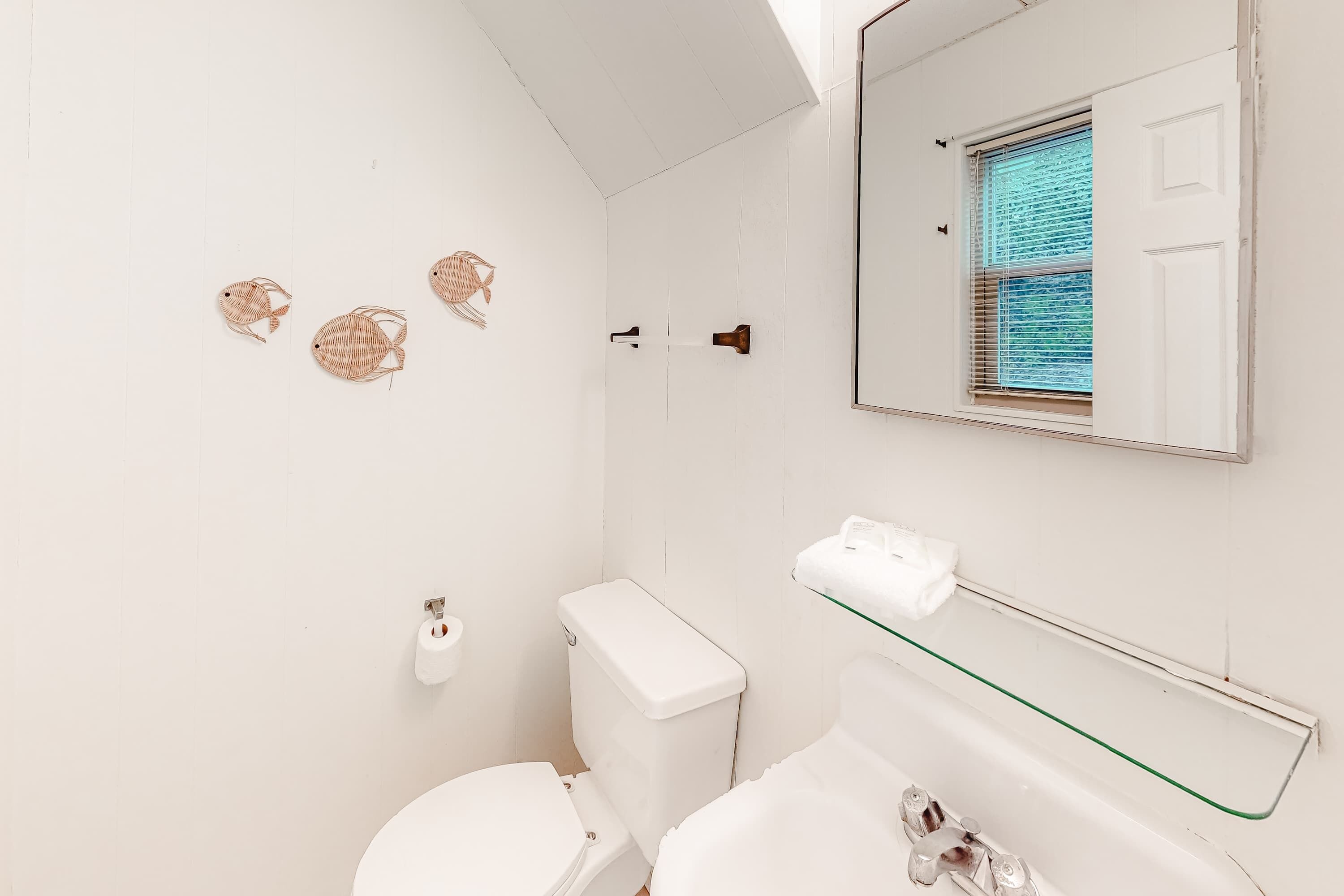 A bathroom with a toilet, a glass shelf, a mirror, and decorative wall art.