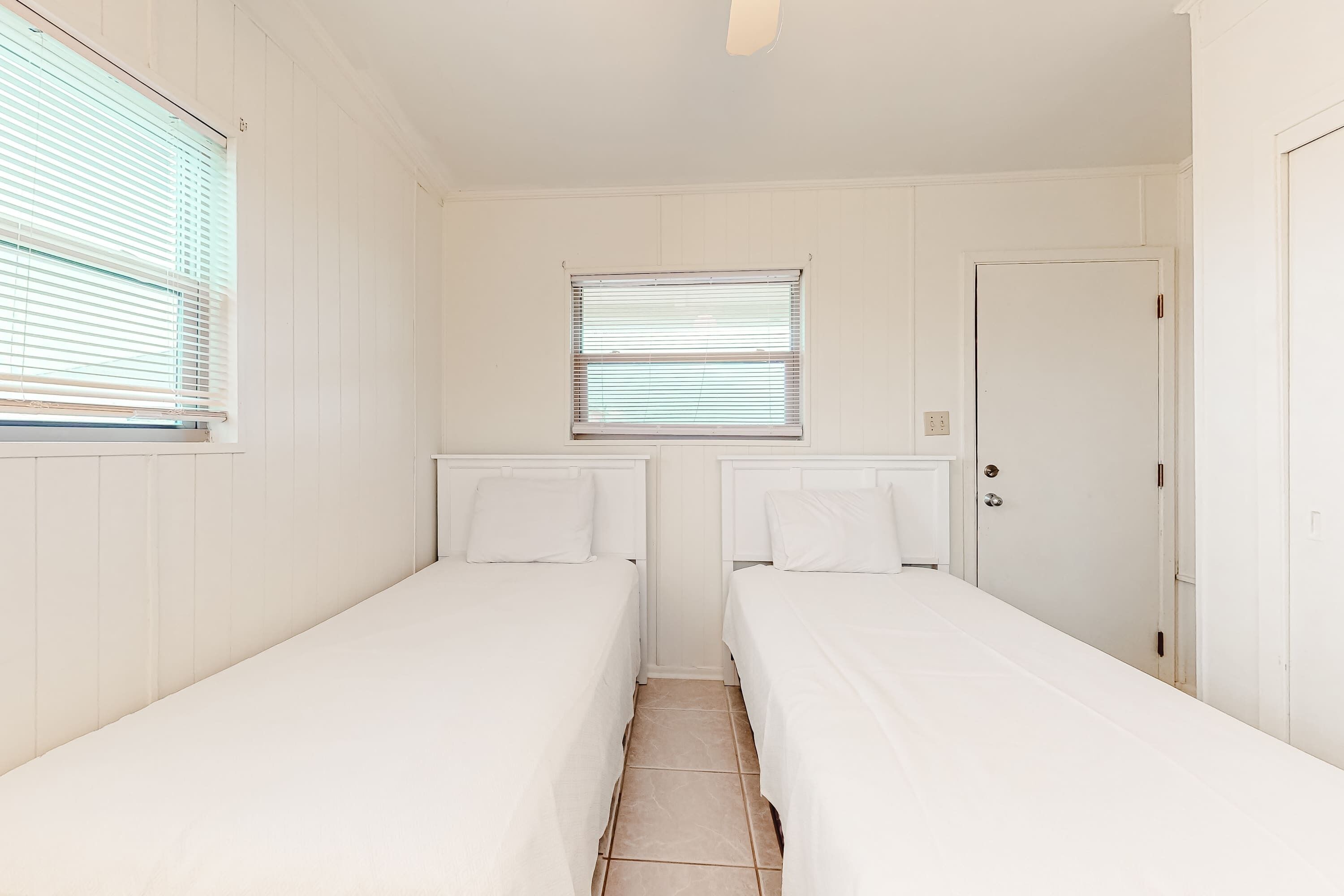 A room with two single beds, a window with blinds, a door, and a tiled floor.