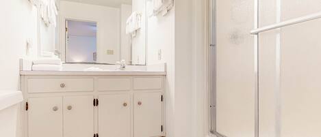 A bathroom with a white vanity, a mirror, a shower, and a door leading to another room.