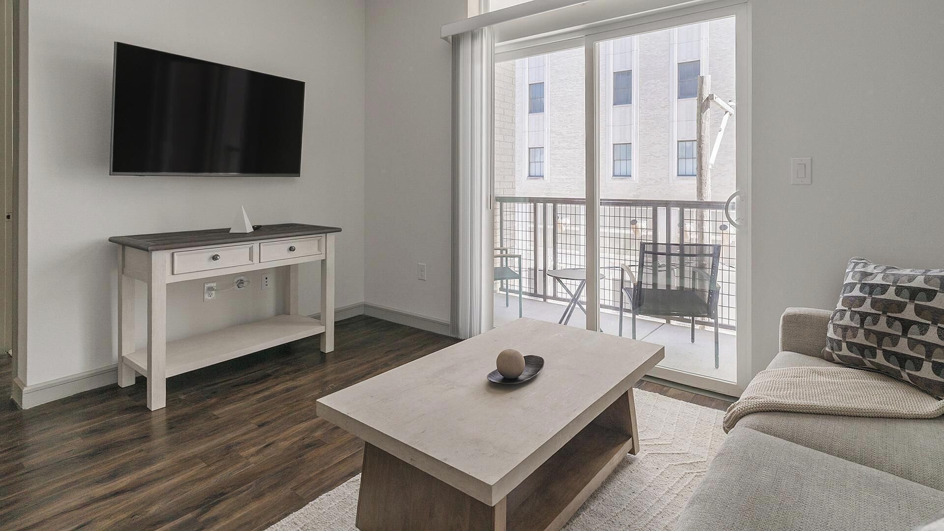 Una sala de estar moderna con una televisión de pantalla plana, una consola blanca, una mesa de centro y un balcón con mobiliario exterior.