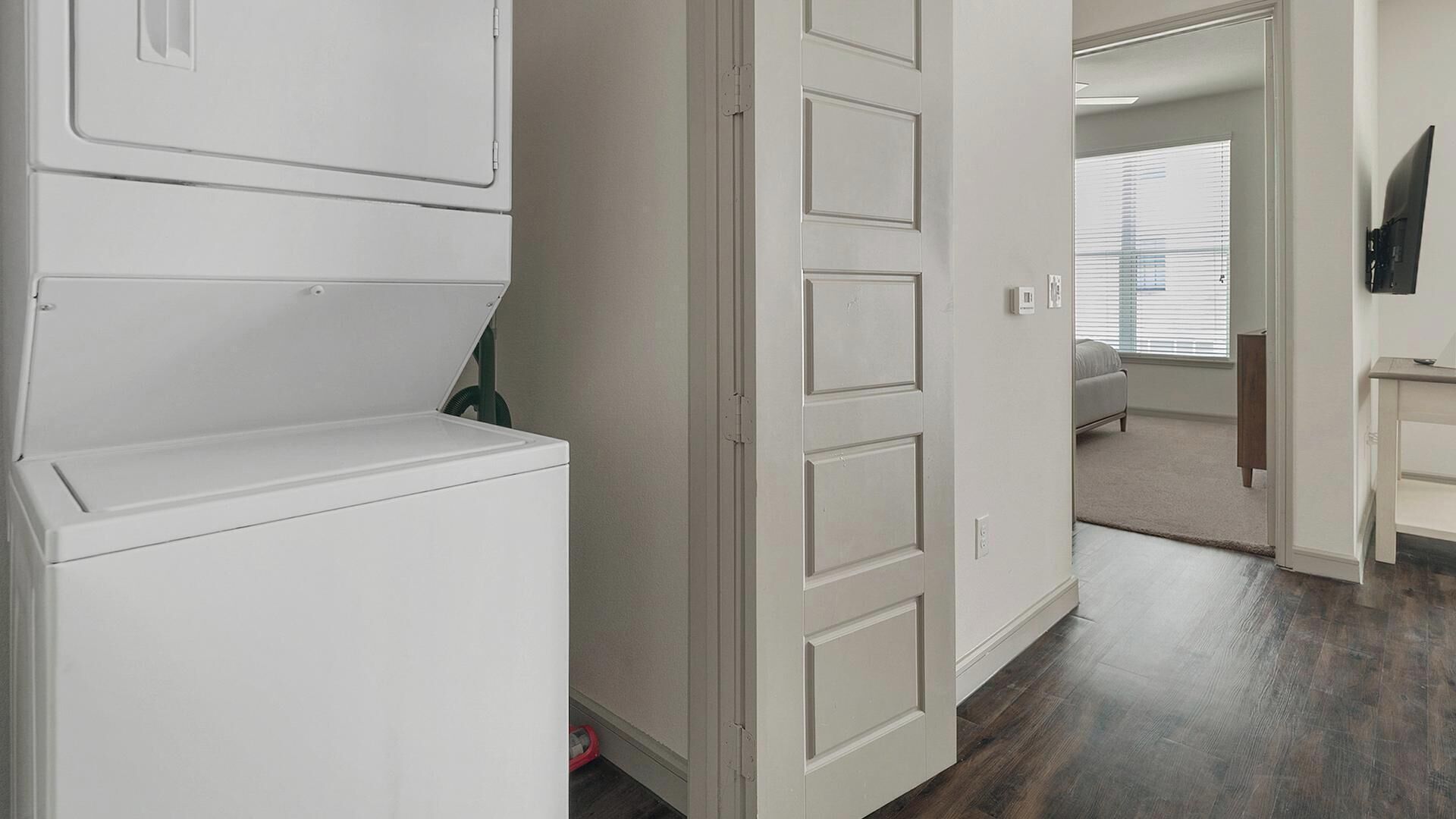 A laundry room with a washer and dryer, a door leading to a bedroom, and a TV mounted on the wall.