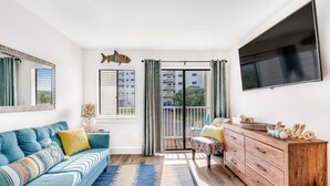Una sala de estar con sofá, cómoda de madera, televisor y balcón con cortinas.