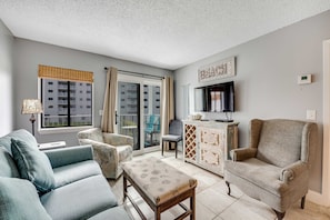 A living room with a sofa, armchair, ottoman, TV, and a cabinet. - Gulf view condo with balcony & shared pools, gym - beach nearby (Fort Morgan)
