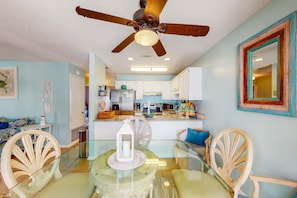A dining area with a glass table, chairs, a ceiling fan, and a kitchen in the background. - Stunning water-view condo with pool - steps away from the beach (Gulf Shores)
