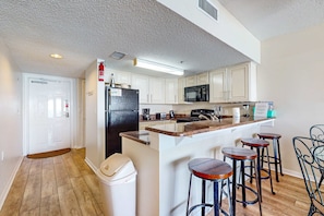 A kitchen with white cabinets, a black refrigerator, a microwave, a coffee maker, and a sink. There are three stools and a trash can. - Tropical Winds beachfront beauty with pool, balcony, grills - close to beach (Gulf Shores)