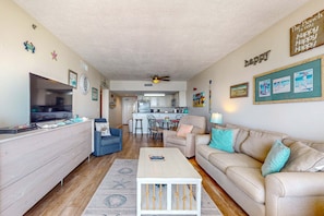 A living room with a TV, sofa, coffee table, and kitchen in the background. - Tropical Winds beachfront beauty with pool, balcony, grills - close to beach (Gulf Shores)
