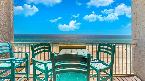 Ein Balkon mit Blick auf Strand und Meer, ein Holztisch und grüne Stühle.