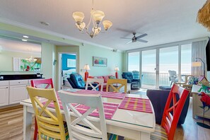 A dining area with a glass table, colorful chairs, and a view of the ocean. - Colorful beachfront condo with Gulf views, pool, sauna, gym & W/D (Gulf Shores)
