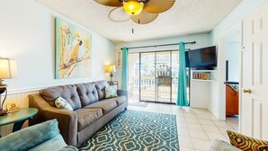 A living room with a sofa, patterned rug, ceiling fan, artwork, and a television. - Home with partial ocean view, central AC, washer/dryer & pool (Gulf Shores)