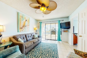 A living room with a sofa, patterned rug, ceiling fan, artwork, and a television. - Home with partial ocean view, central AC, washer/dryer & pool (Gulf Shores)