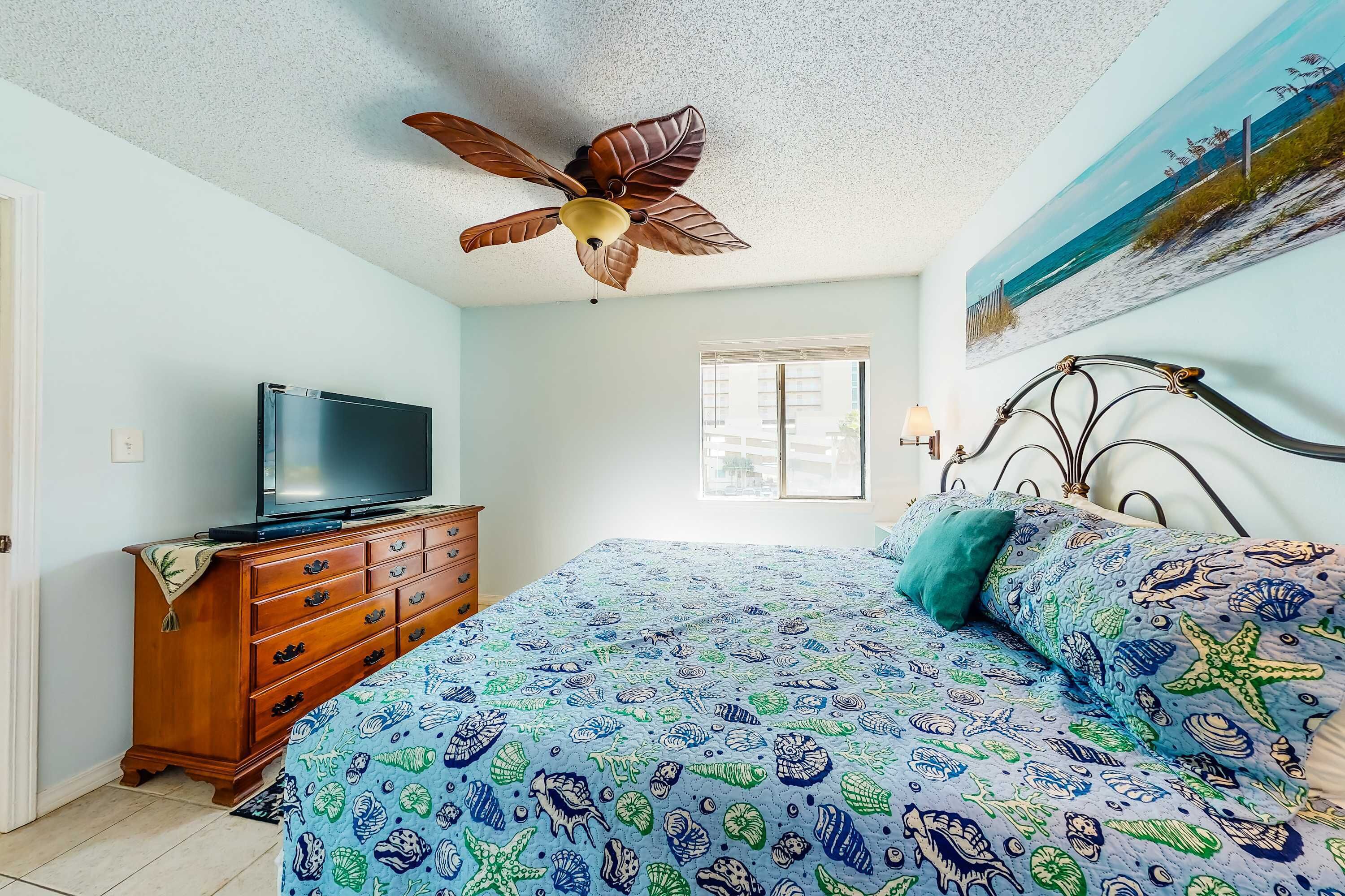 Ein Schlafzimmer mit Bett, Deckenventilator, Kommode, Fernseher und Fenster.