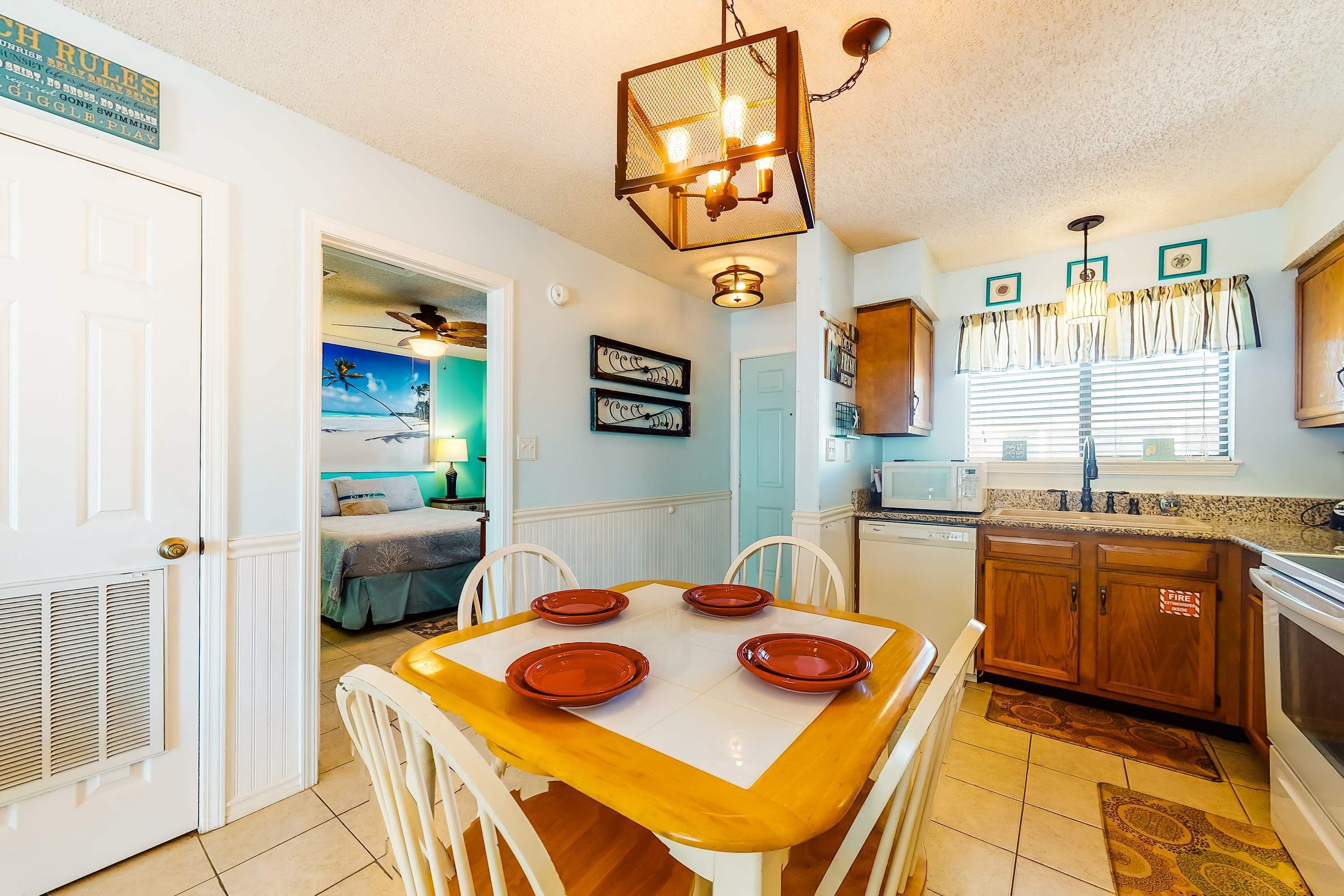 A dining area with a wooden table, four chairs, a chandelier, and a kitchen with wooden cabinets and a microwave.