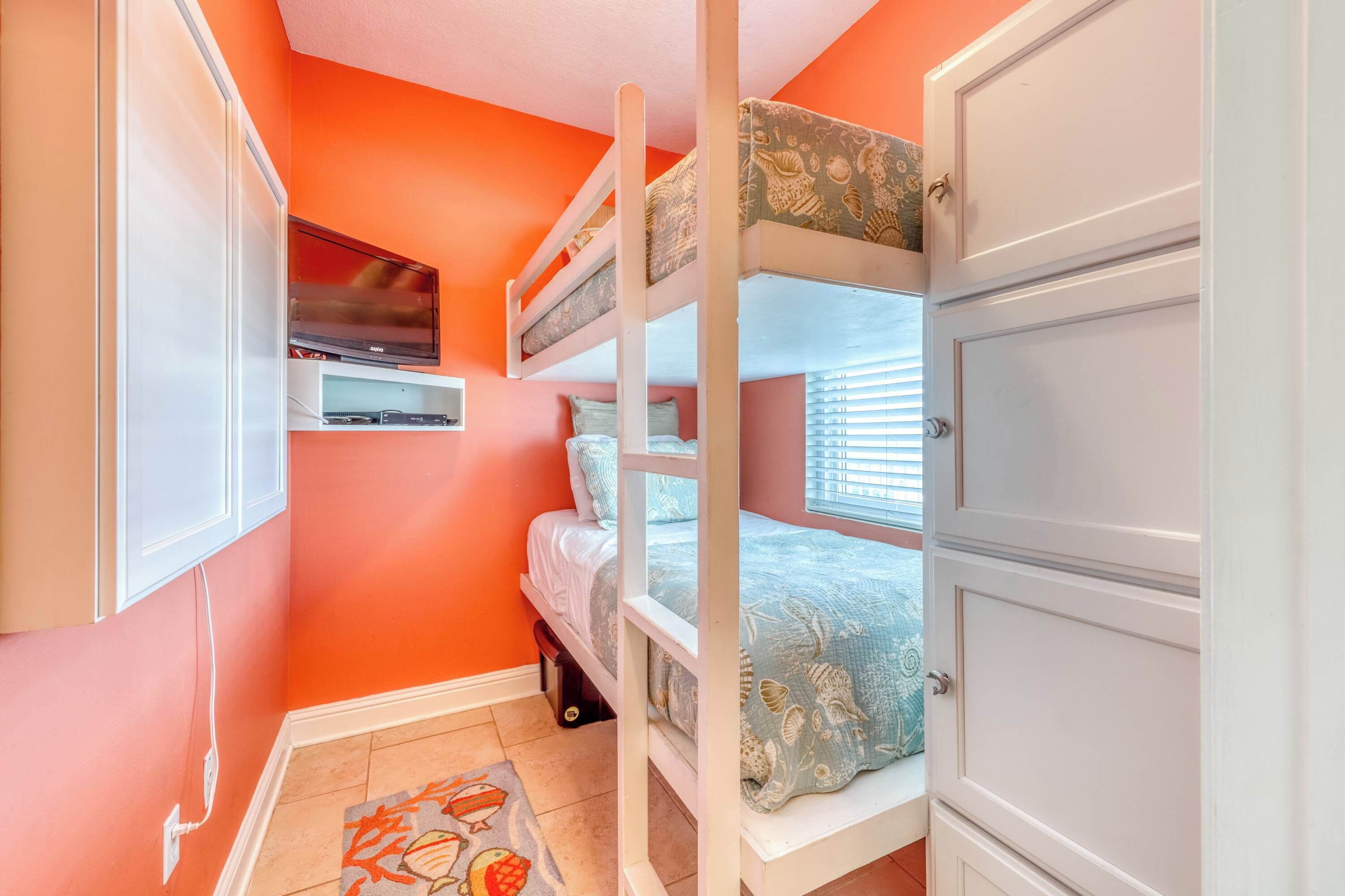 Une chambre avec un lit superposé, une télévision posée sur une étagère, une armoire et une fenêtre avec des stores.