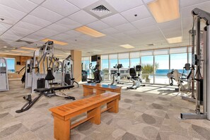 A gym with exercise equipment, a wooden bench, and large windows with a view. - Gulf front condo w swim-through outdoor & indoor pool, hot tub, and sauna (Gulf Shores)