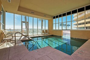 An indoor swimming pool with clear water, large windows, and a view of palm trees and a building. - Gulf front condo w swim-through outdoor & indoor pool, hot tub, and sauna (Gulf Shores)