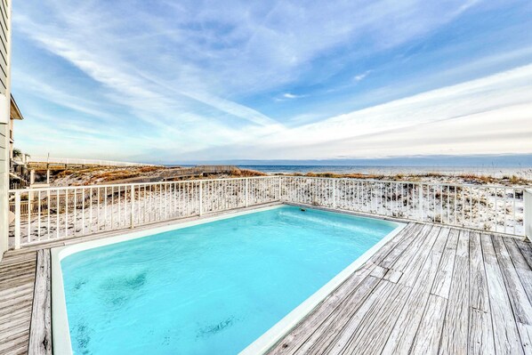 Una piscina rectangular con plataforma de madera, rodeada por una valla blanca, con vista a una playa y al océano.