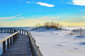 Ein hölzerner Steg führt zu einem Strand mit Dünen und blauem Himmel.