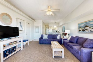 Ein Wohnzimmer mit Fernseher, Sofas, einem Couchtisch, einem Deckenventilator und einer Küche im Hintergrund.