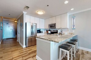 A modern kitchen with white cabinetry, stainless steel appliances, a granite countertop, and a bar stool seating area. - Upgraded beachfront condo w balcony & resort pools, hot tubs & sauna (Gulf Shores)