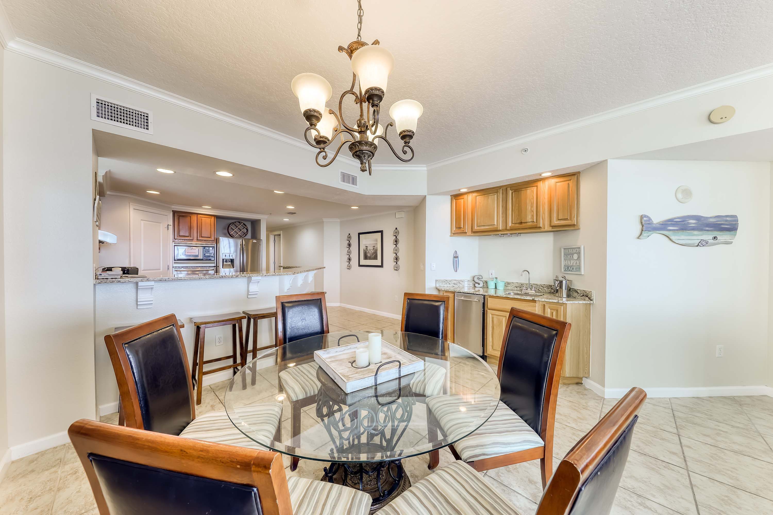 A dining area with a glass table surrounded by chairs, a chandelier, and a kitchen in the background.