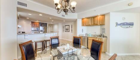 A dining area with a glass table surrounded by chairs, a chandelier, and a kitchen in the background.