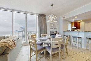 A dining area with a round table, chairs, and a view of the city through large windows. - Luxurious beachfront condo w incredible Gulf view & shared pool hot tub (Gulf Shores)