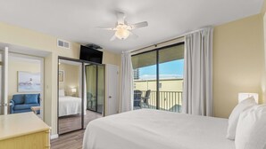 A hotel room with a bed, a desk, a chair, a balcony with curtains, and a ceiling fan. - Beachfront condo with balcony overlooking Gulf of Mexico & resort pool (Gulf Shores)